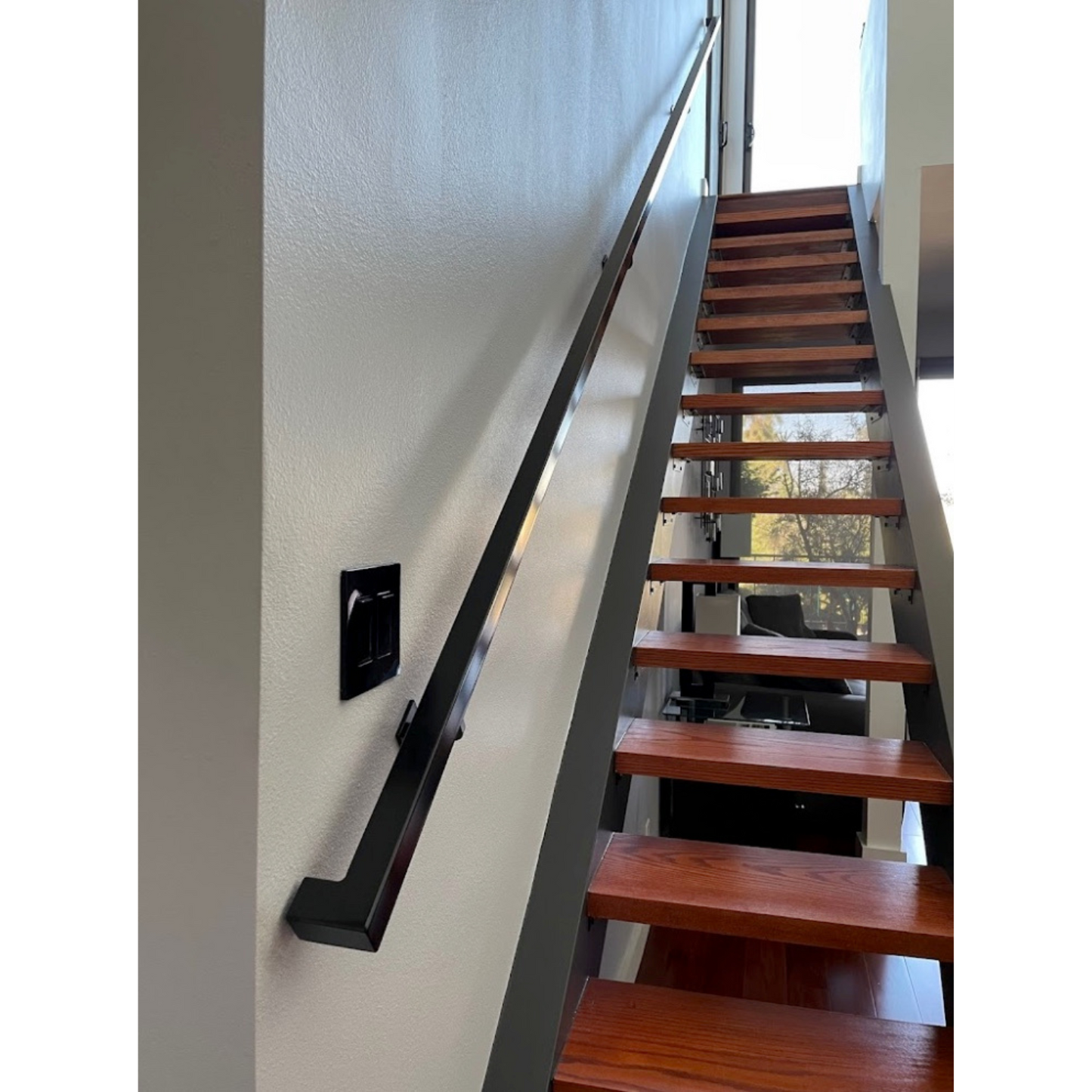 1.5"x1.5" Metal Handrail mounted on a white wall above floating, wooden steps with large, glass windows on the upper and lower levels in the background