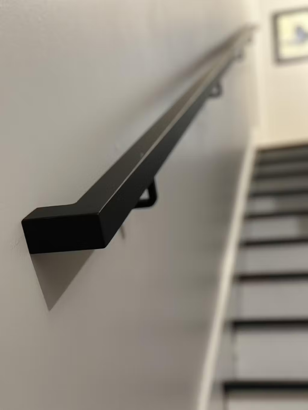 close view of the 1.5"x1.5" Metal Handrail on light grey wall above steps with black tread and white risers